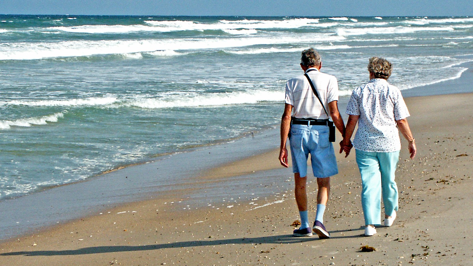 Healthy ageing: an older couple walking hand in hand along the beach at the water's edge